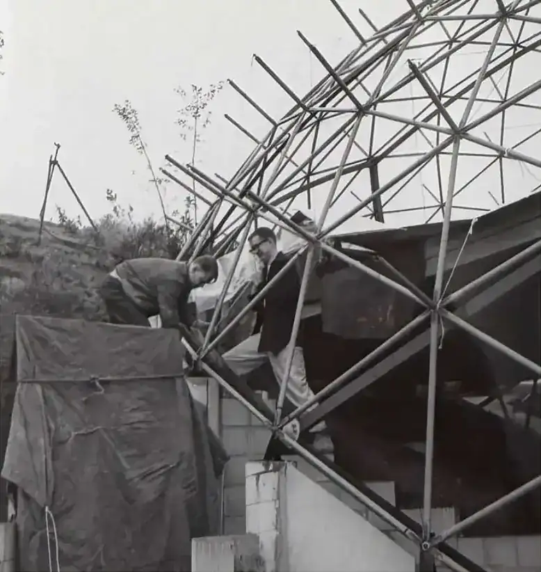 The geodesic frame rising over a block wall, two men at its base